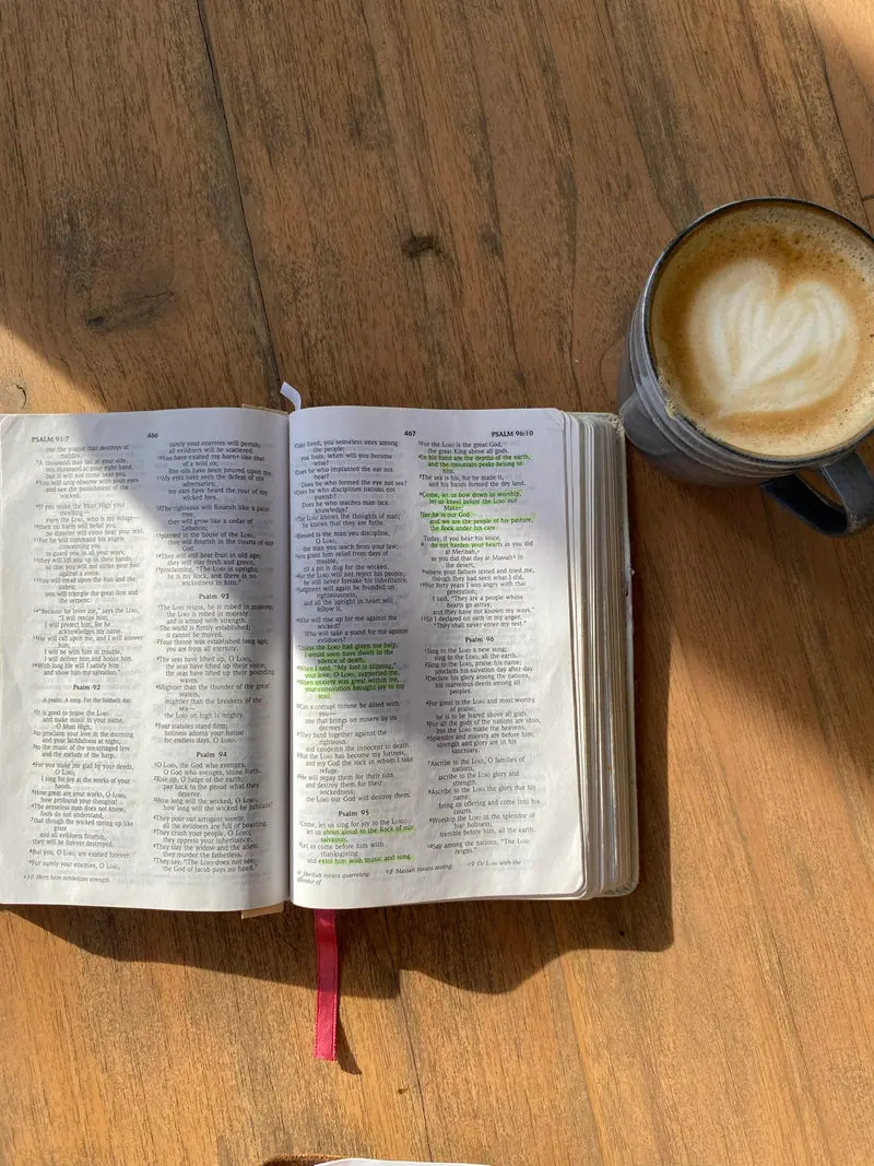 A coffee beside an open Bible on a wooden table in morning light